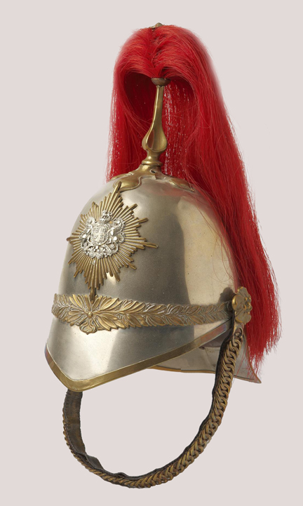 The helmet, circa 1880, is of white metal construction. Across the front seam a decorative gilt band of olive leaves and a band of oak leaves conceals the rear seam. A plain gilt chain backed with leather and attached to the helmet with gilt rosettes. The plume is attached to the helmet through a gilt spike resting on a plain quatrefoil.